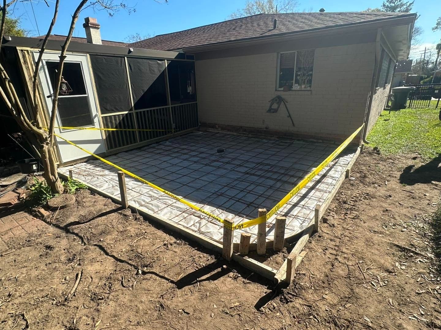 Backyard patio slab under construction beside a house, with yellow safety tape and dirt around it