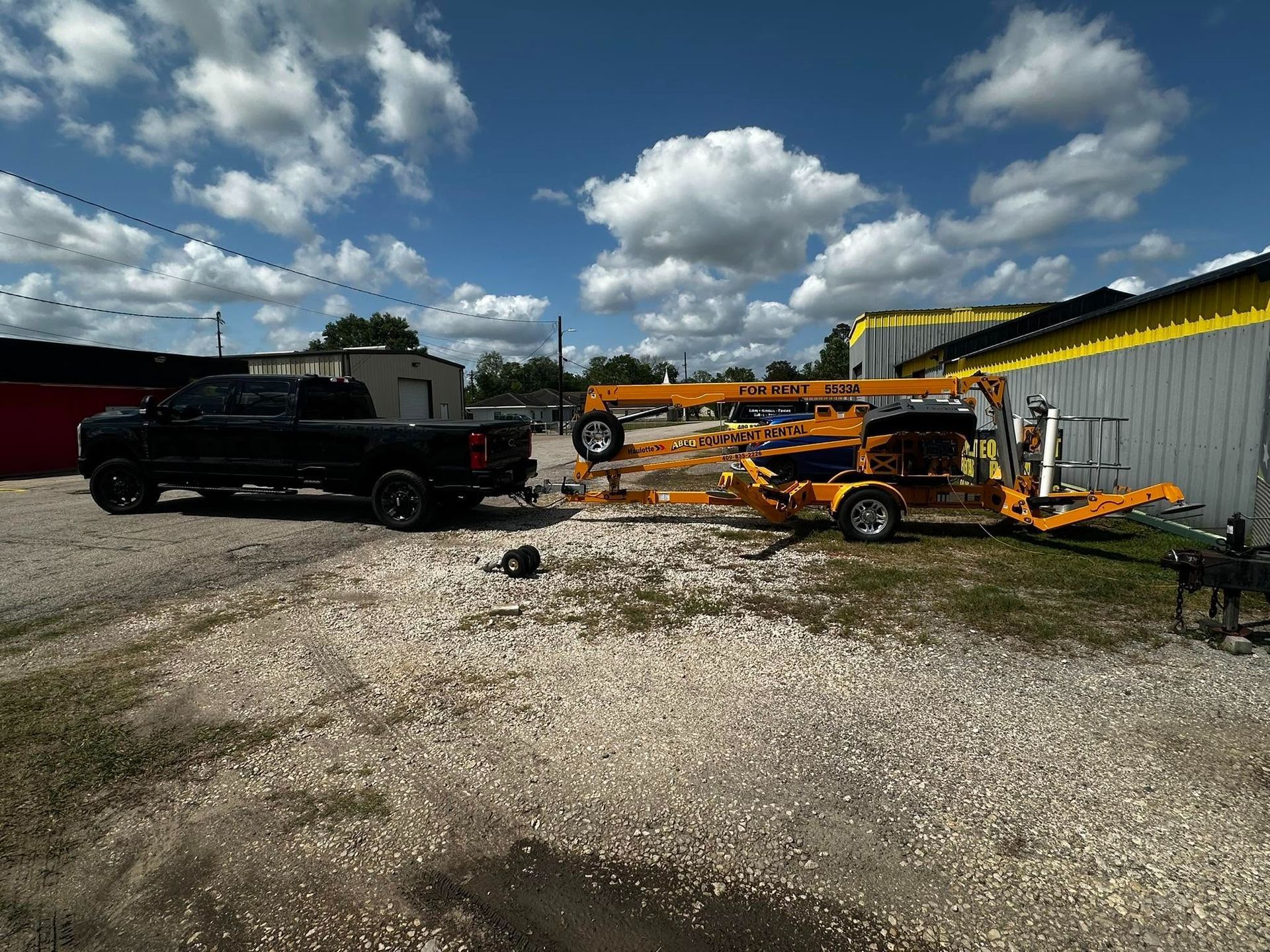 Black pickup and orange equipment parked on gravel beside a yellow-roofed building under a cloudy sky
