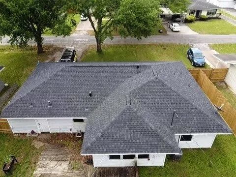 Aerial view of a gray-roofed house with a driveway, yard, and trees nearby