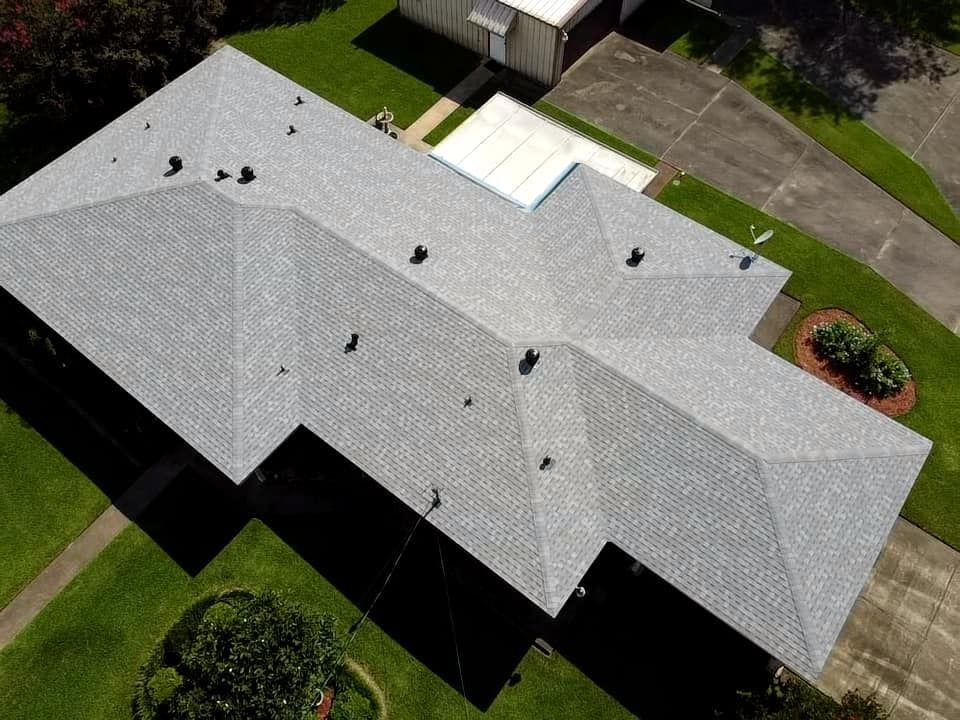 Aerial view of a gray-roofed house with a driveway, lawn, and surrounding trees