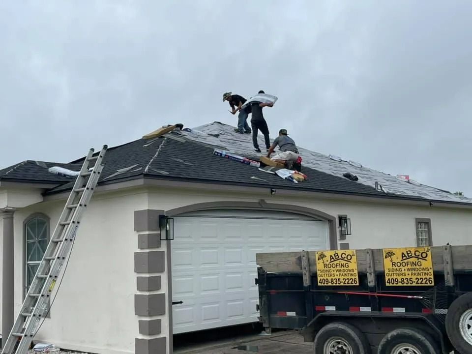 Workers repairing a shingled roof beside a ladder and dump trailer on a house garage