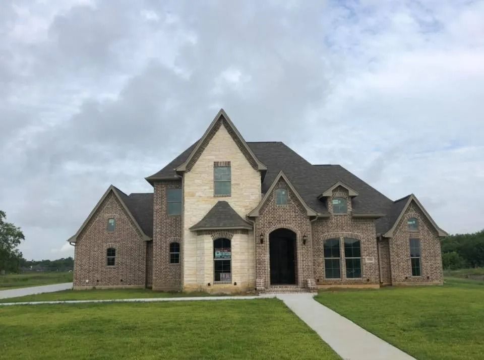 Large stone suburban house with multiple gables, arched entry, and a front lawn under cloudy skies