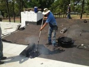 Worker in a white hat applying hot tar to a flat roof with a long tool.