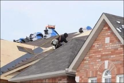 Roofers installing shingles on a house roof with blue tarps and a brick gable.