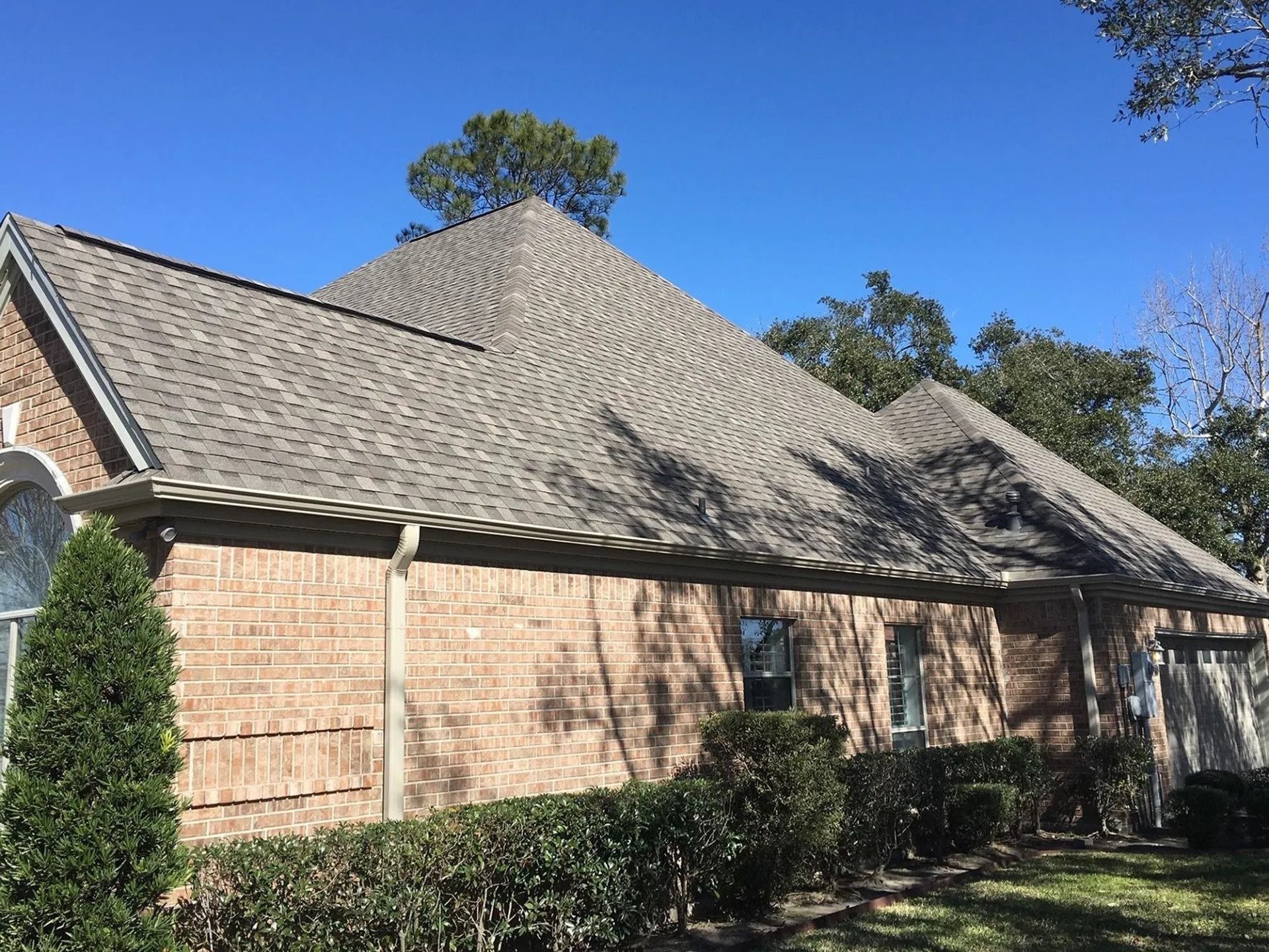 Brick house with a steep gray roof, bushes in front, and a clear blue sky above.