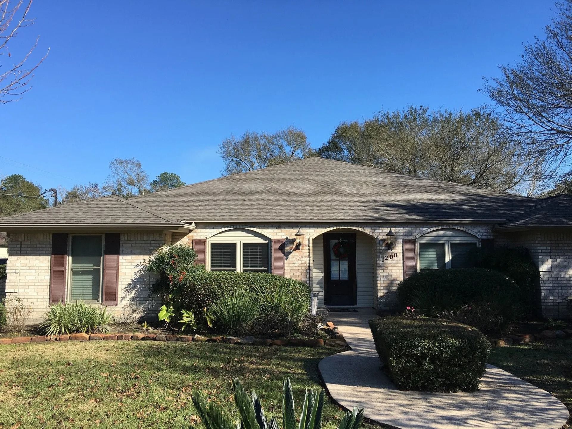 Single-story brick house with a front walkway, shrubs, and a trimmed lawn under a clear blue sky
