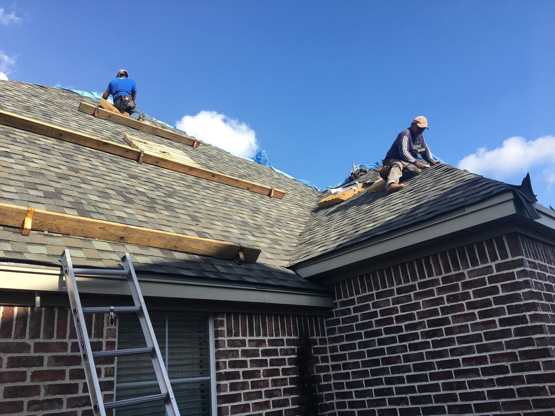 Workers repairing a roof on a brick house, with ladders and blue sky above.