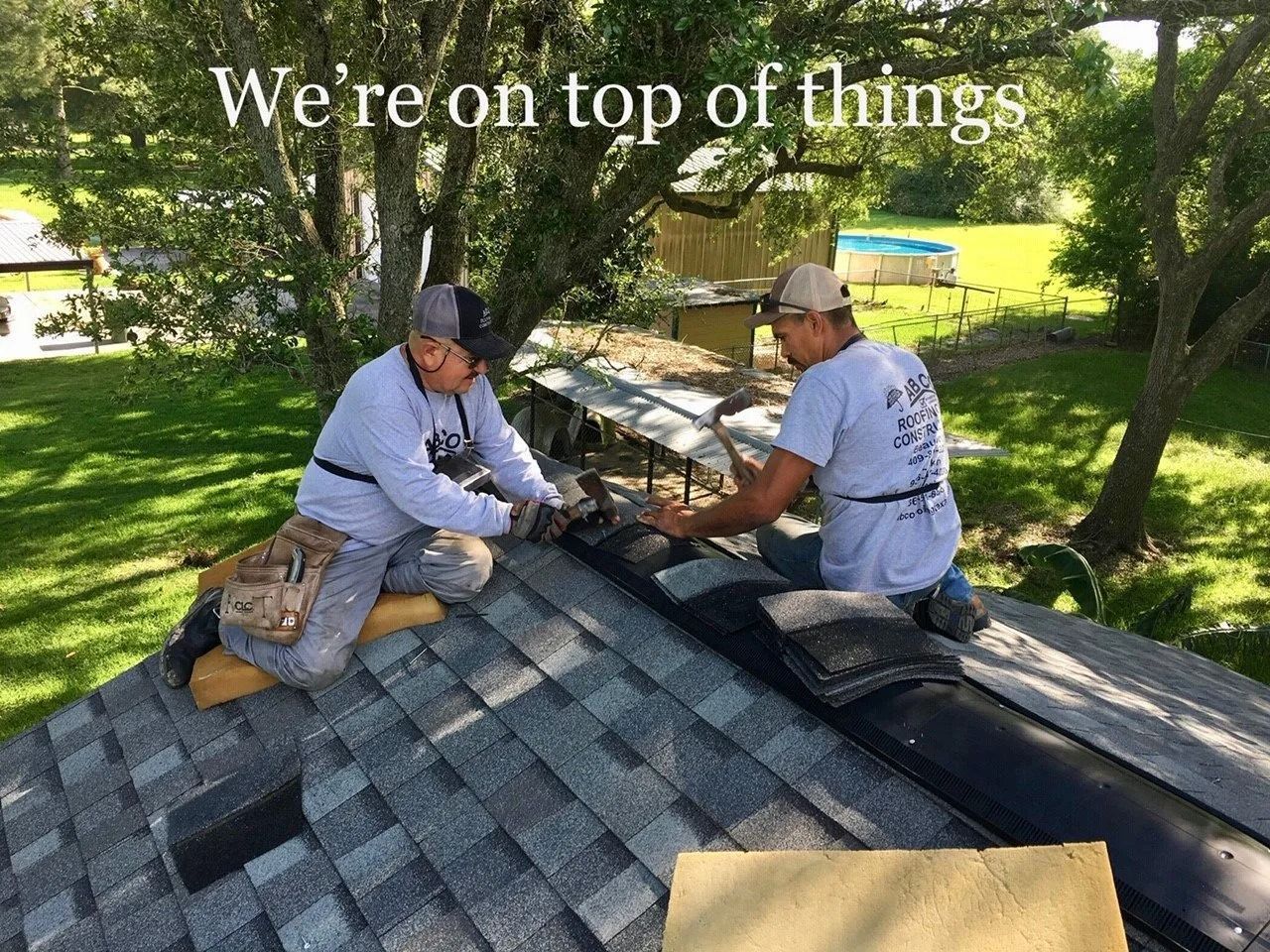 Two workers kneeling on a shingled roof, installing shingles under trees on a sunny day.