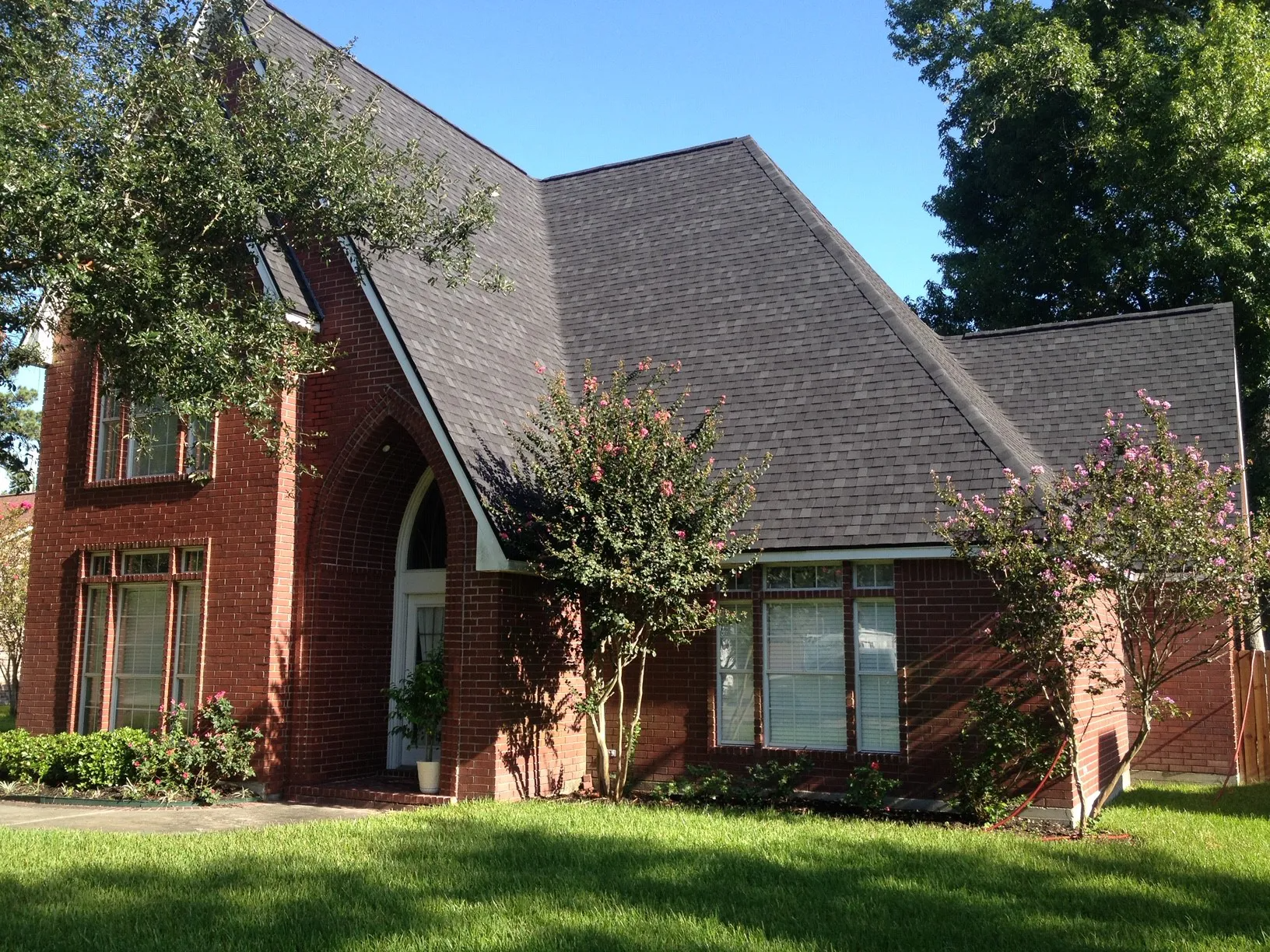Red-brick suburban house with steep gray roof, arched entry, and green lawn under sunny skies