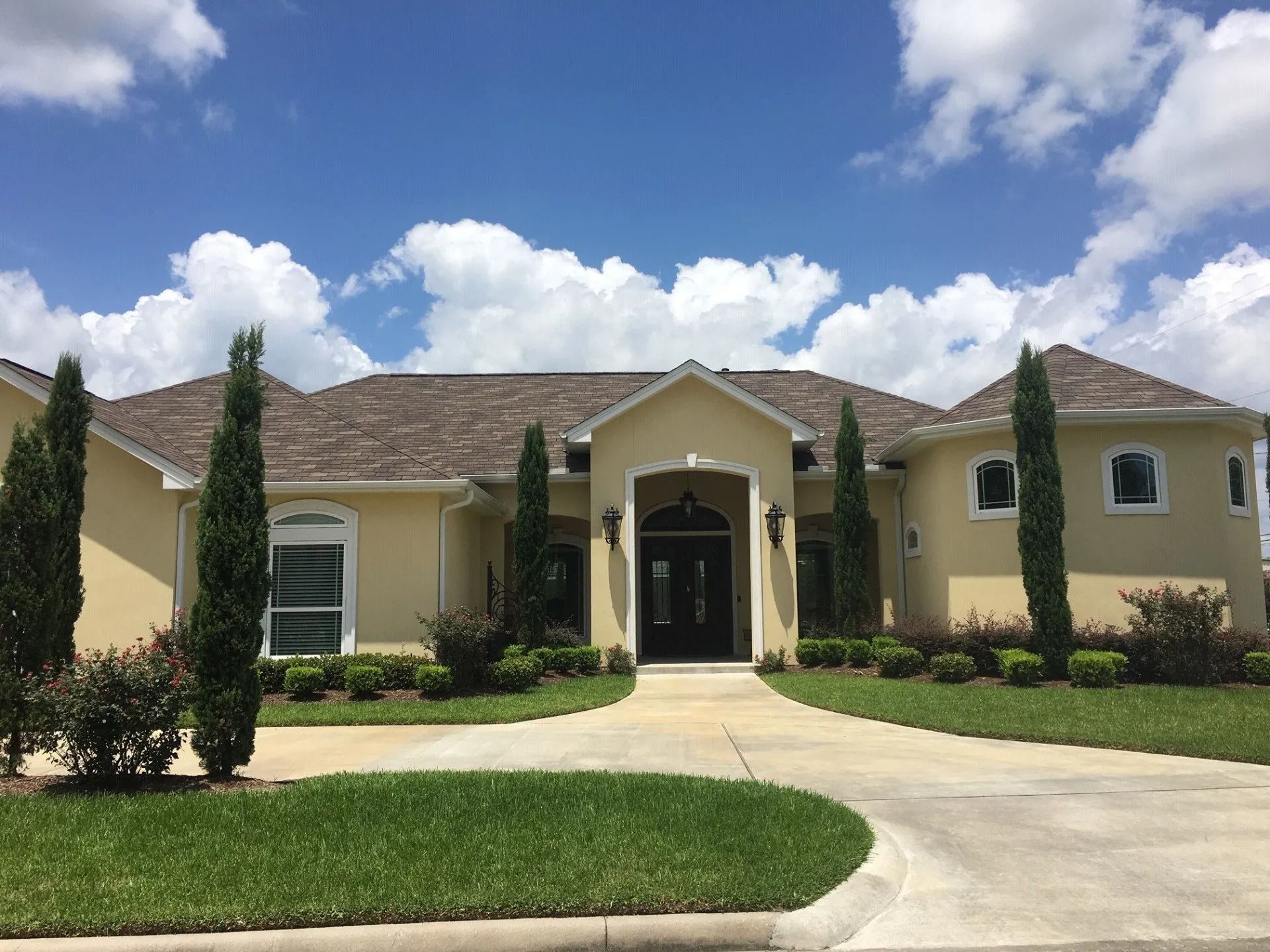 Single-story beige house with a curved driveway, green lawn, and tall shrubs under a blue sky.