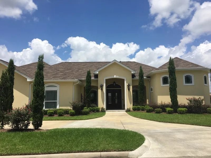 Large beige house with a manicured lawn, curved driveway, and tall cypress trees under a blue sky.