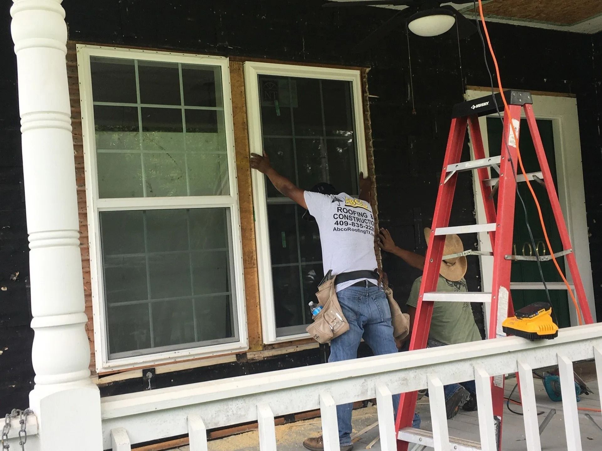Worker painting a house exterior beside a window, with a red ladder on the porch.