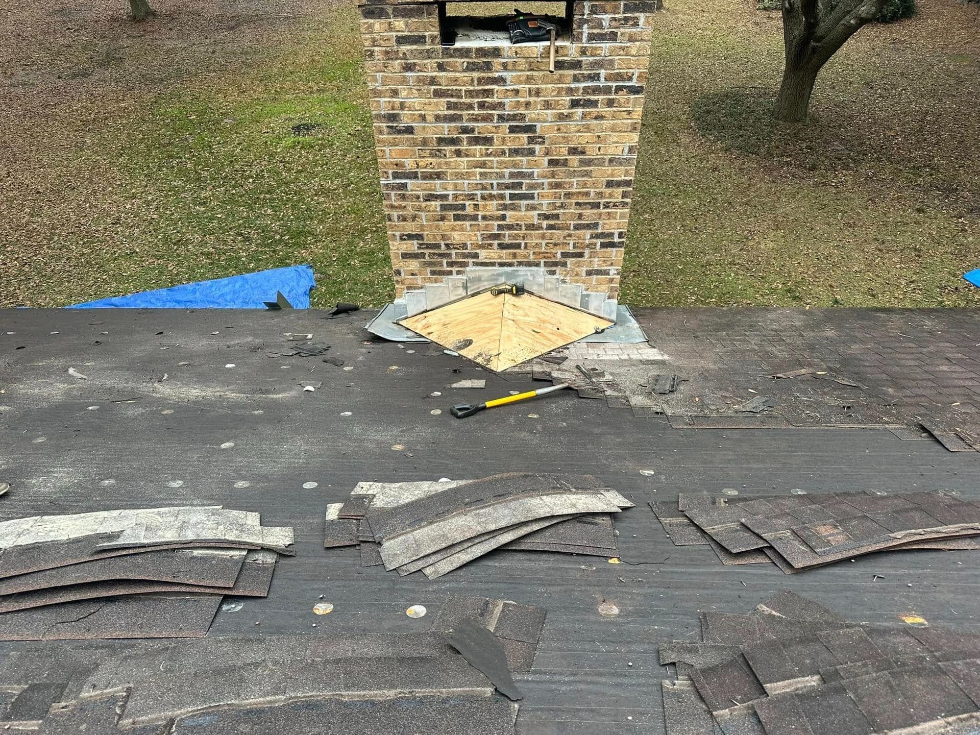 Damaged shingle roof around a brick chimney with exposed underlayment and debris.