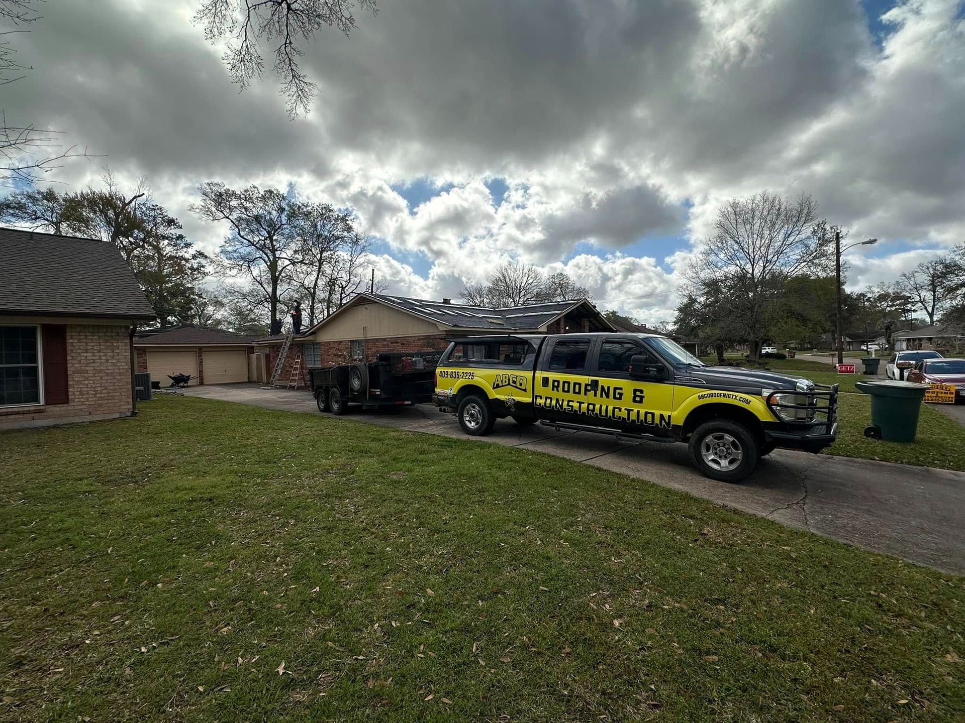 Yellow utility truck parked in a driveway beside a brick house under a cloudy sky