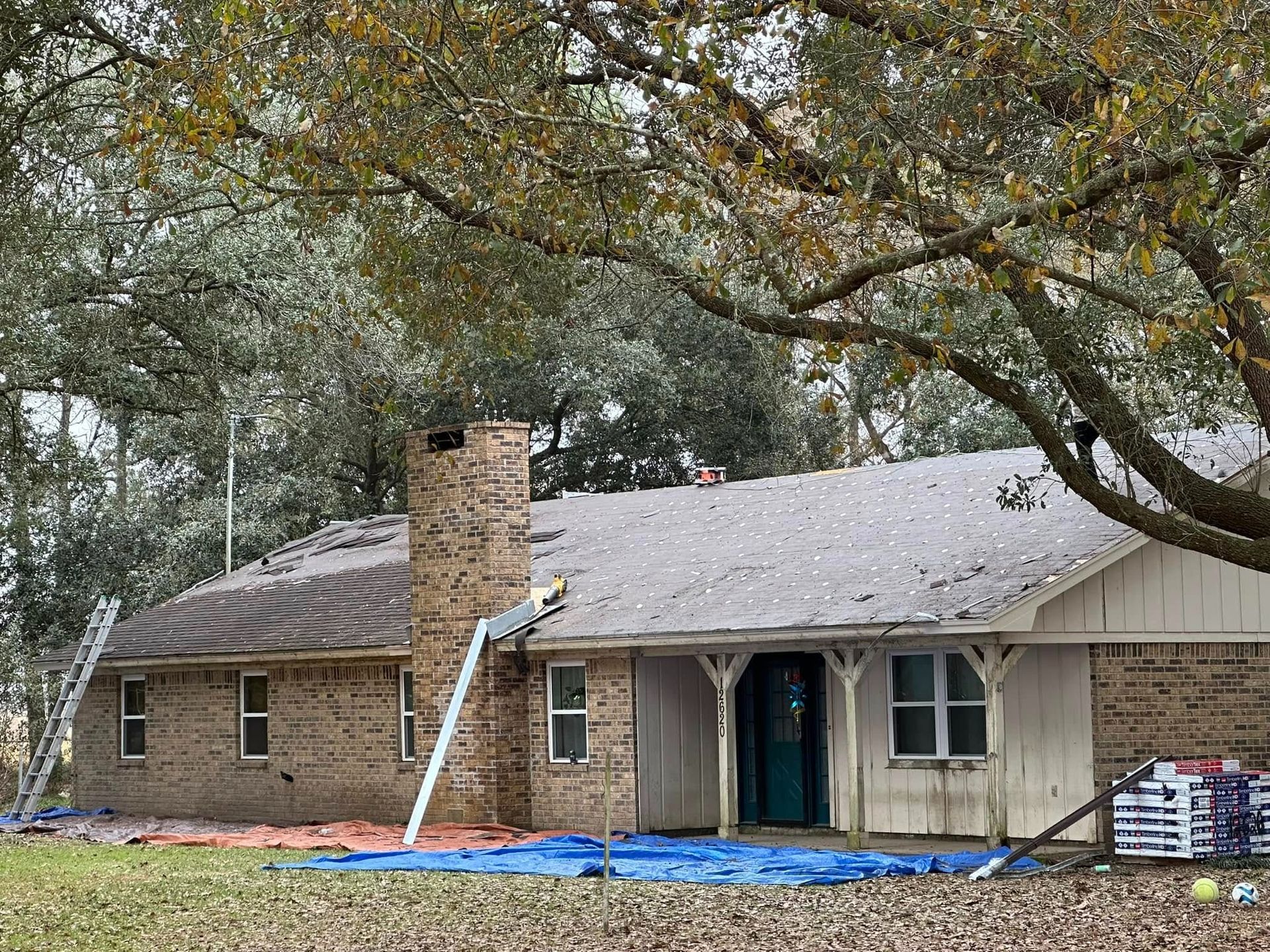 Damaged house with tarp-covered roof and fallen tree limbs in a yard after storm damage