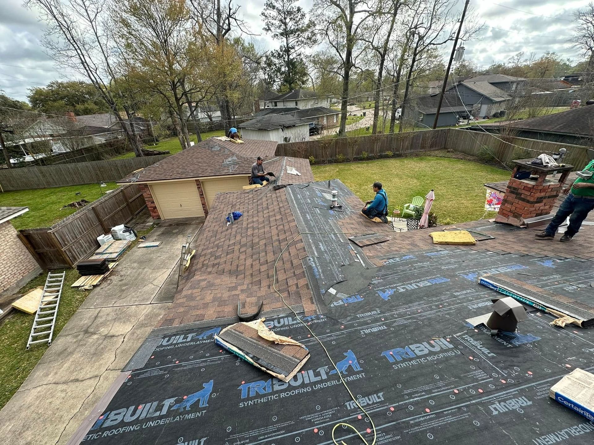 Workers installing roofing underlayment on a steep roof, with ladders and tools in a tree-lined yard