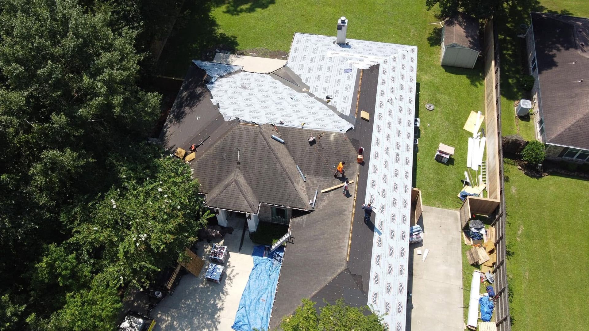 Aerial view of a house with a partially replaced roof and workers on the roof and in the yard.
