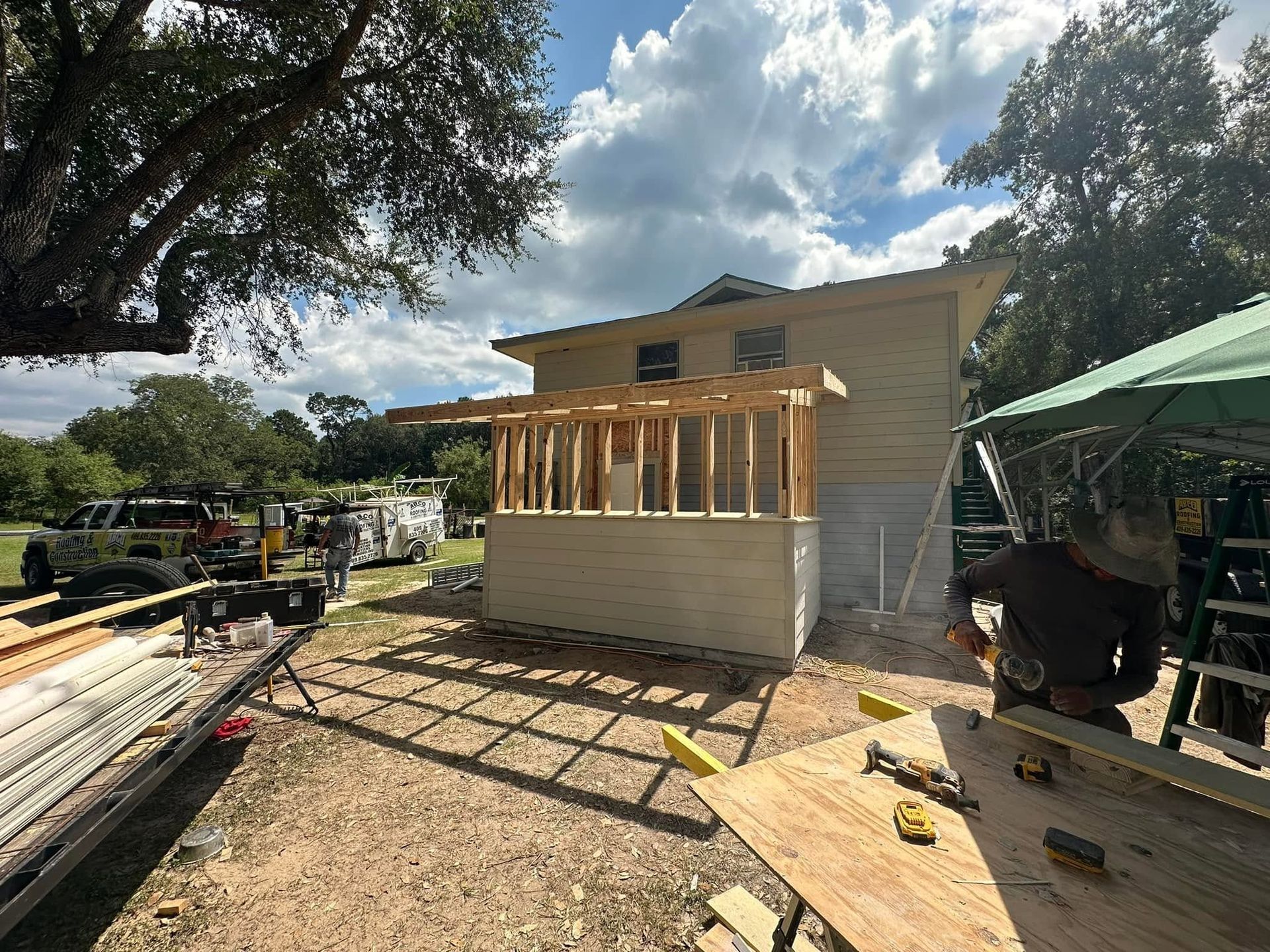 Partially built wooden porch beside a house, with construction materials and a truck in a sunny yard
