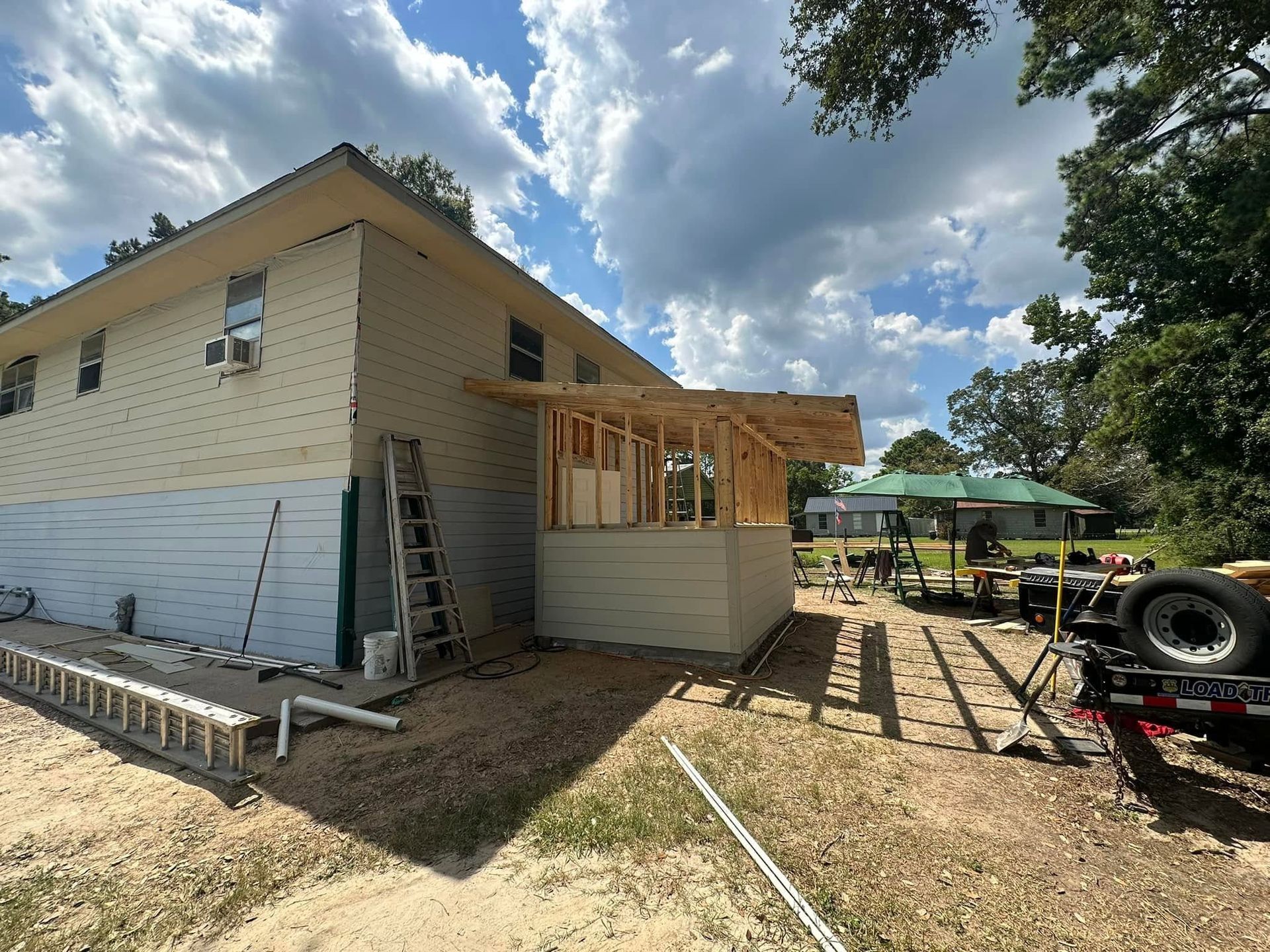 House under construction with ladder, exposed framing, and trailer in a dirt yard under a cloudy sky