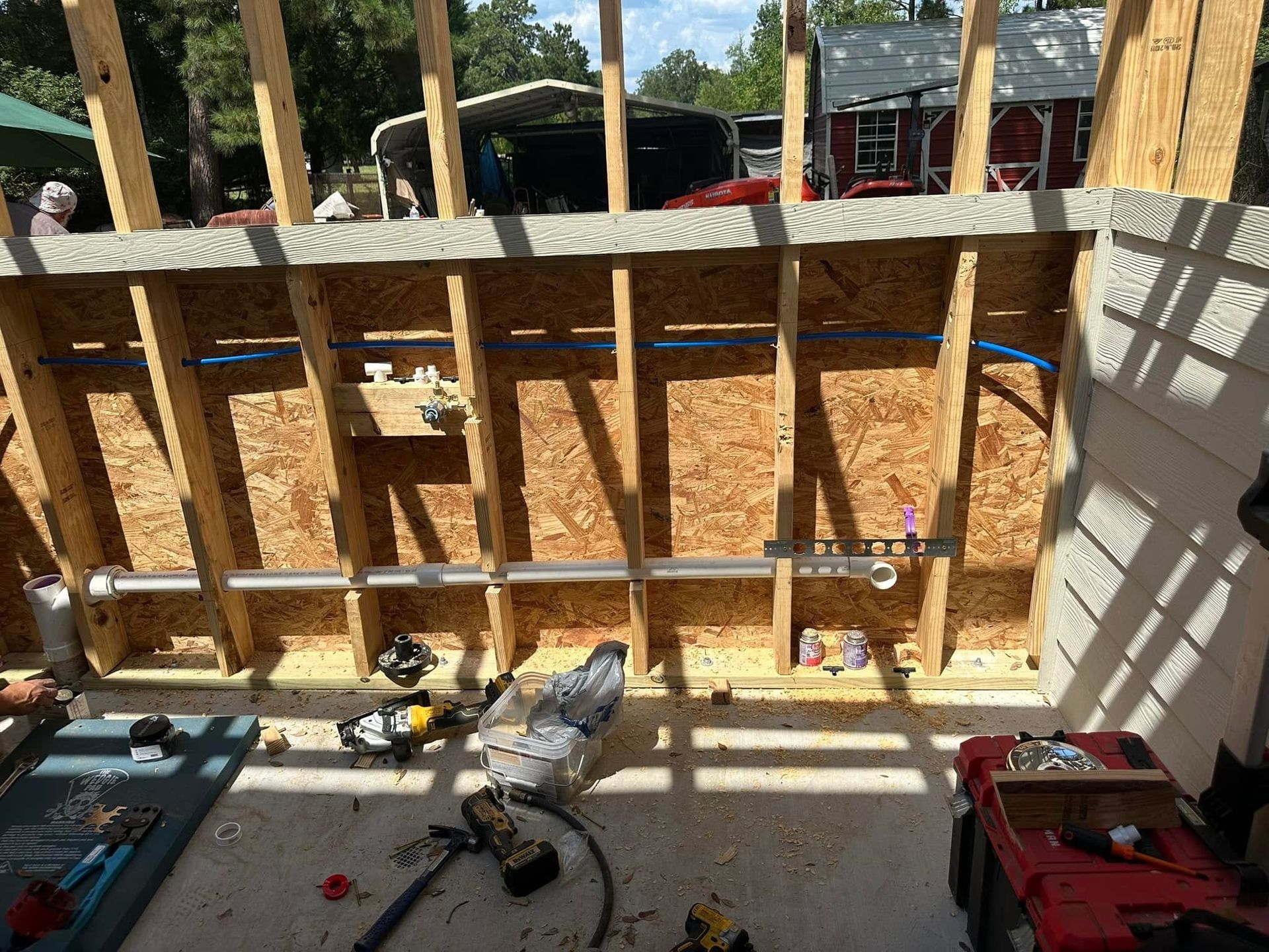 Partially framed wall under construction, with tools on a floor inside a room opening to a yard.