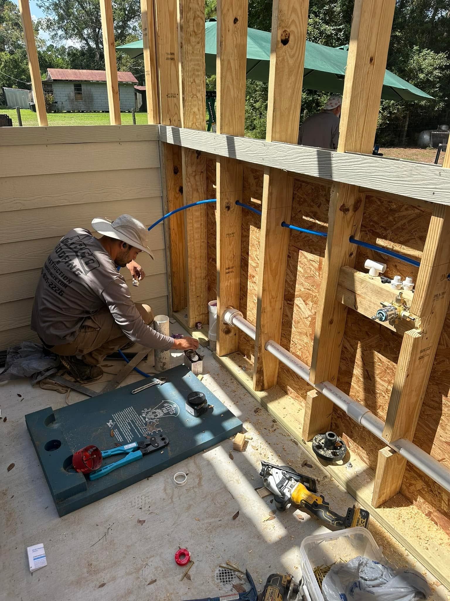Worker kneeling on a porch floor, installing blue conduit inside unfinished wooden wall framing.
