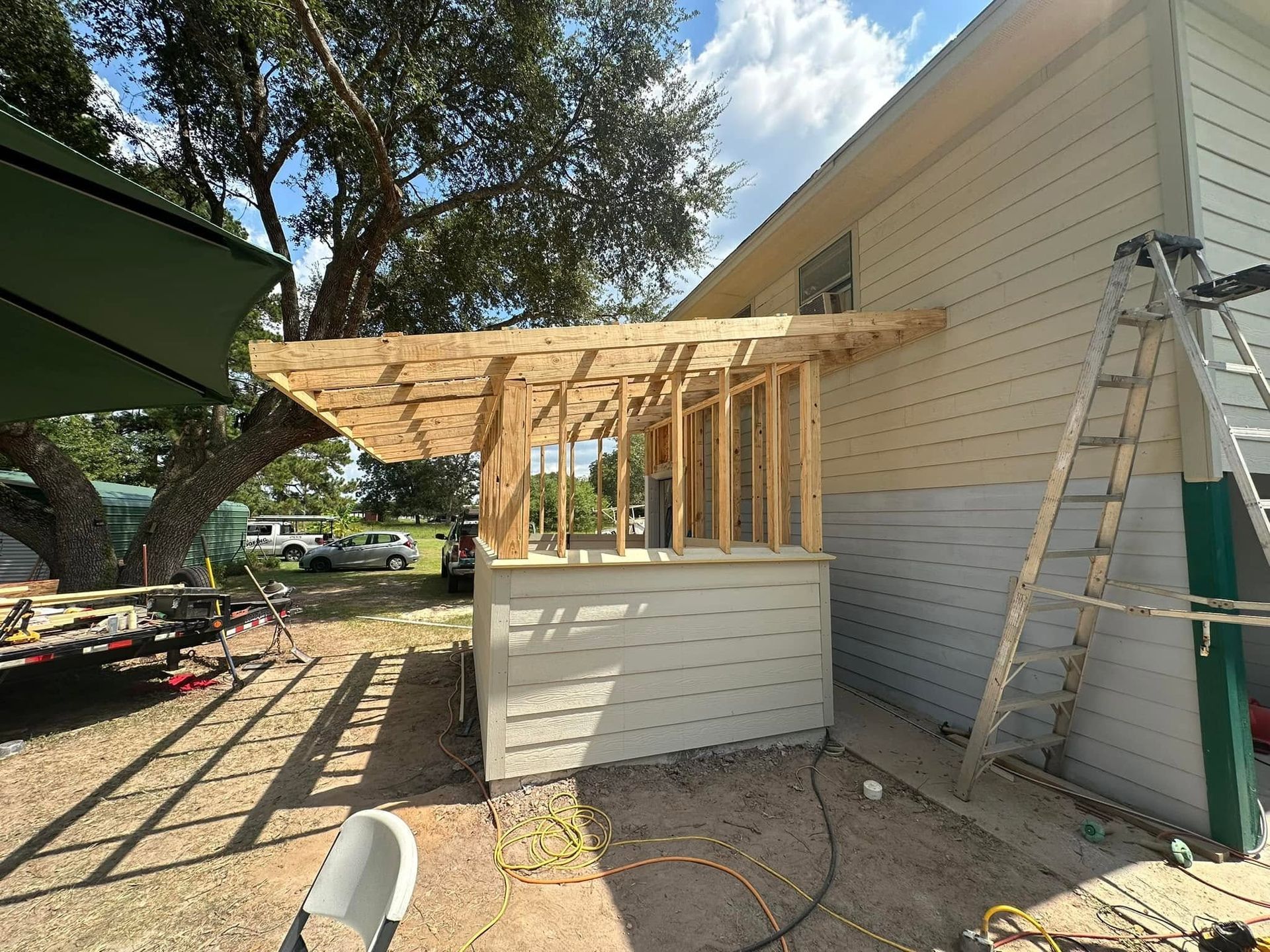 Partially built wooden patio roof beside a house, with a ladder and construction materials nearby