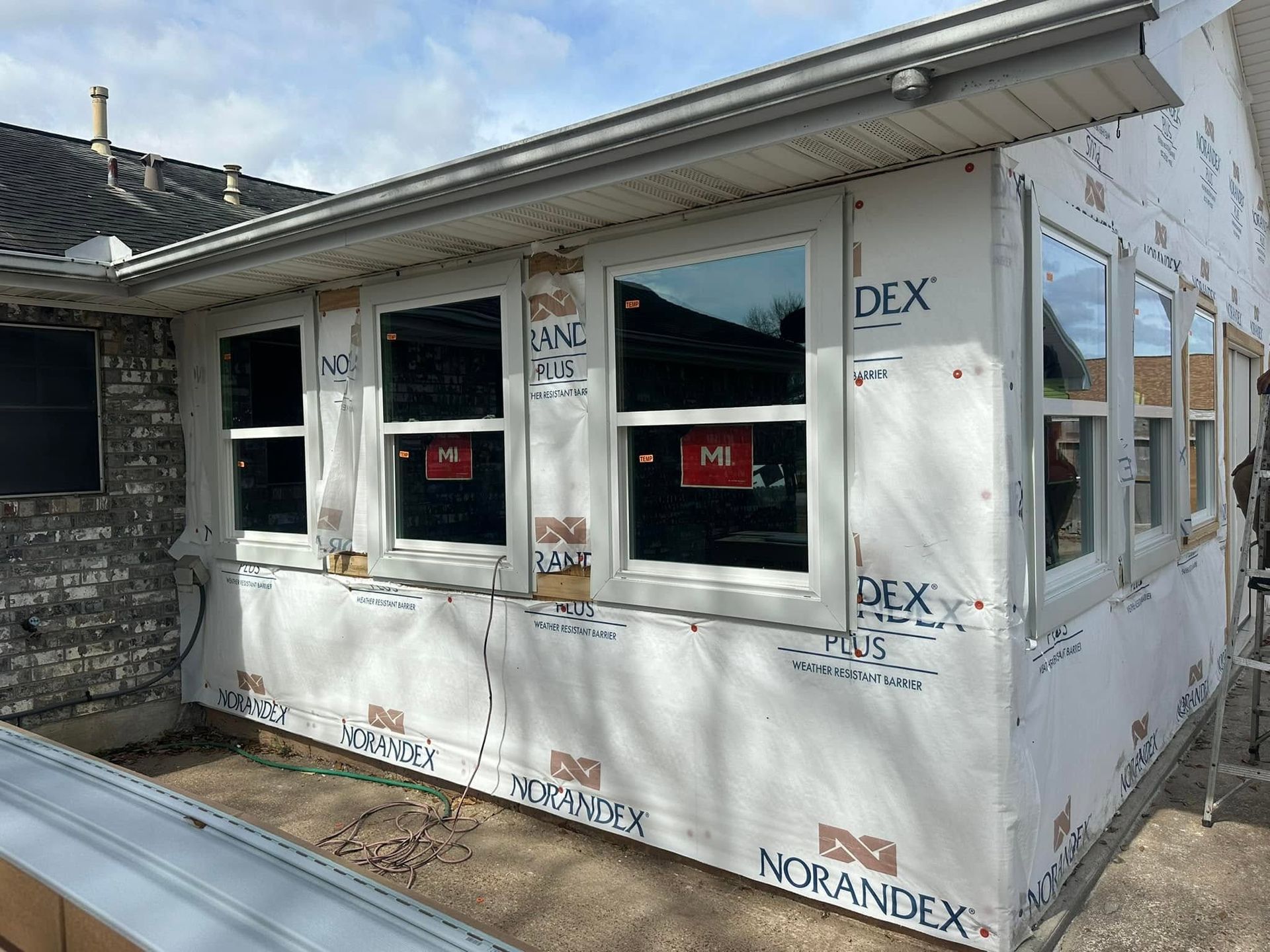 White house extension under construction, wrapped in insulation with windows installed and red safety signs.