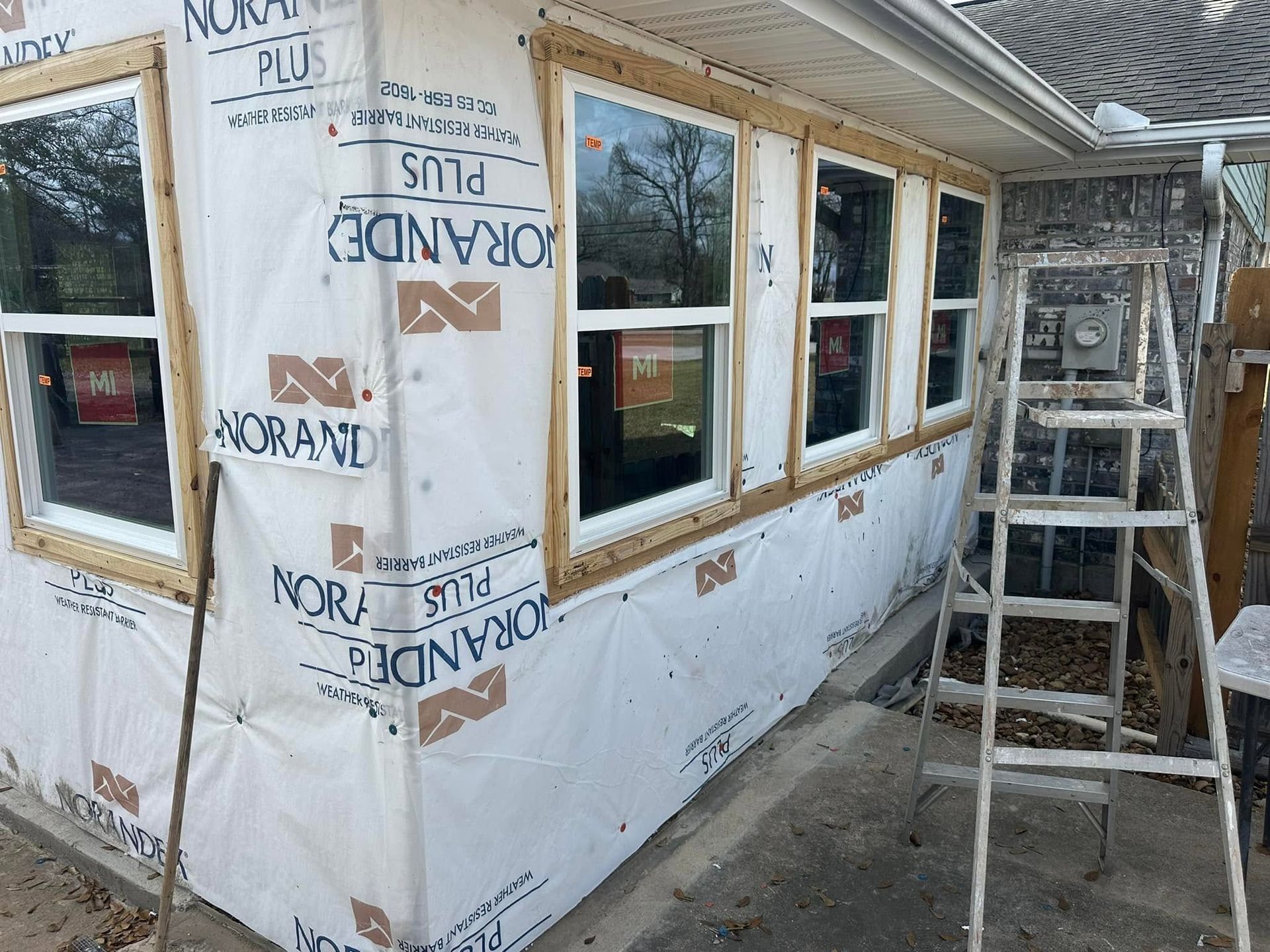 House under construction wrapped in white house wrap, with windows and a ladder beside the wall.
