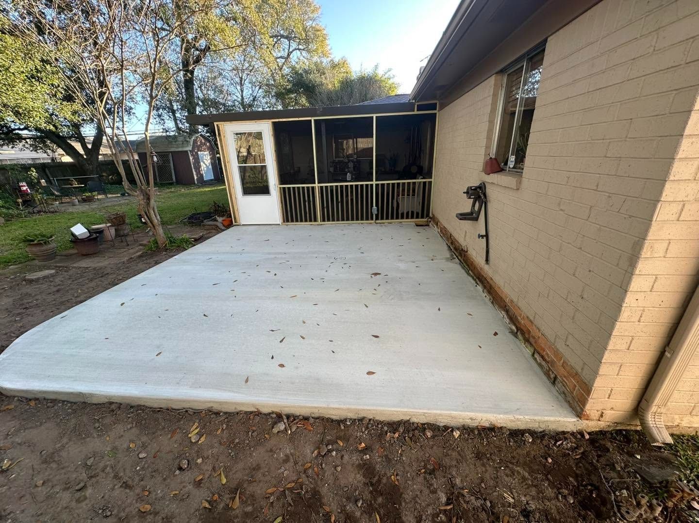 Concrete patio beside a tan brick house, with a small screened porch and yard trees.