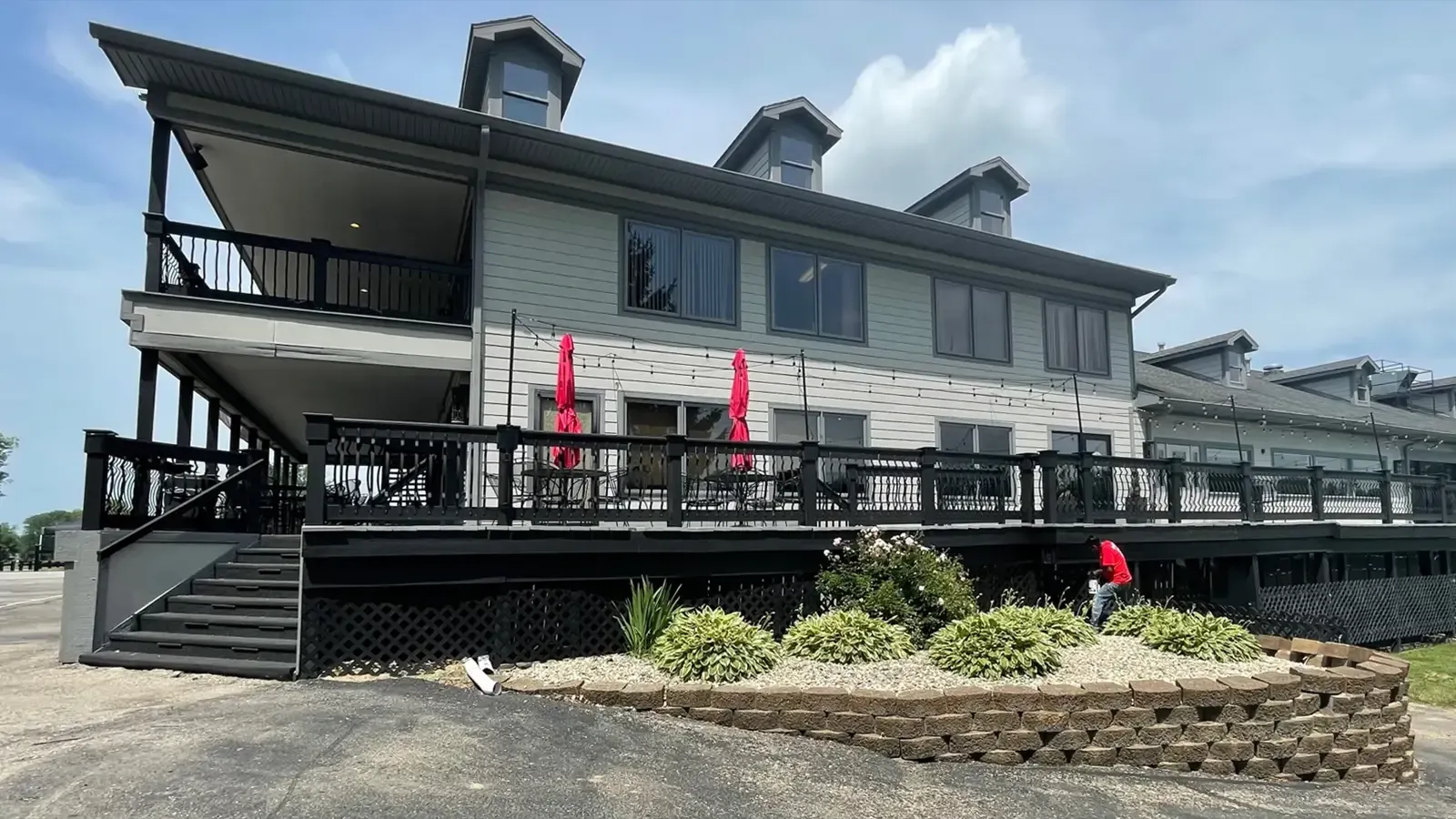 Two-story building with a deck, black railing, gray siding, and red umbrellas. A person is working near landscaping.