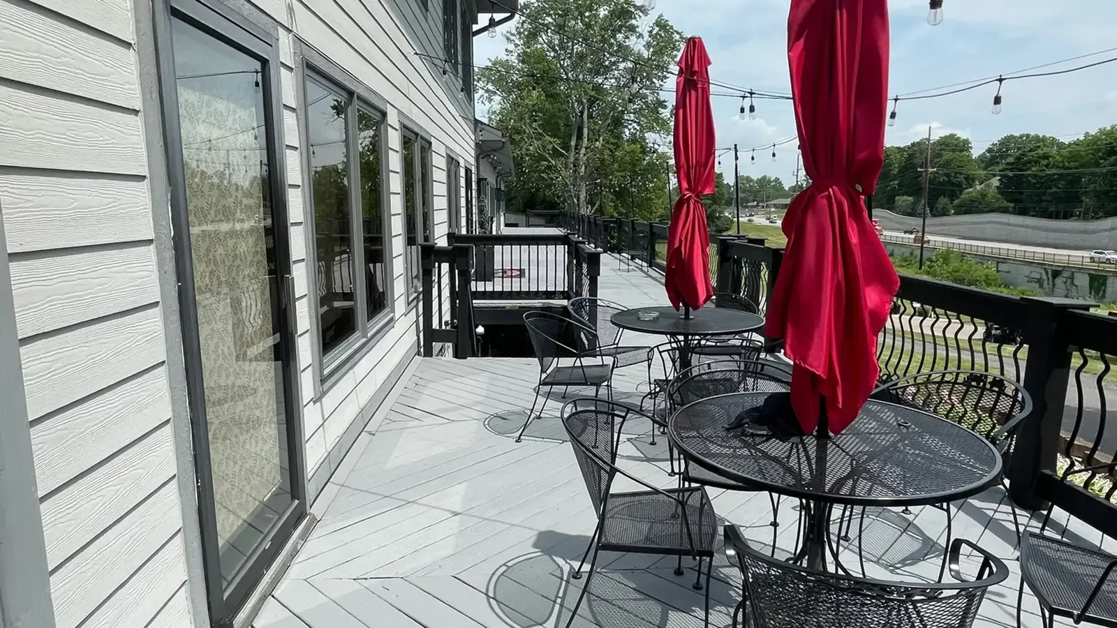 Outdoor restaurant patio with red umbrellas, tables, and chairs. Grey siding and black railings.