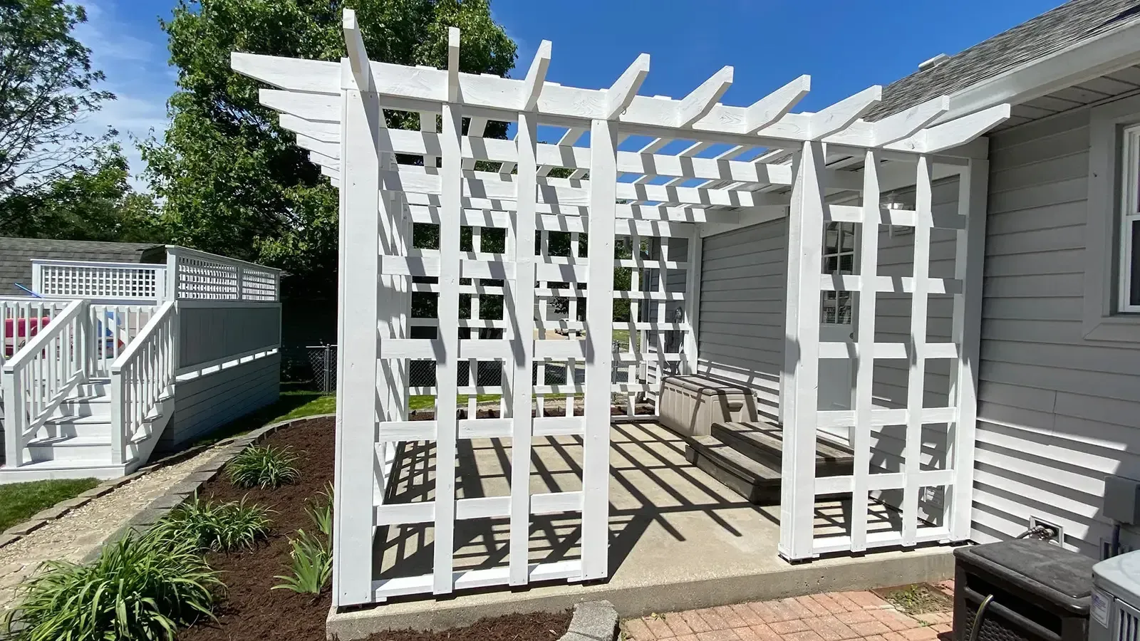 White lattice pergola next to a house with a bench, surrounded by landscaping.