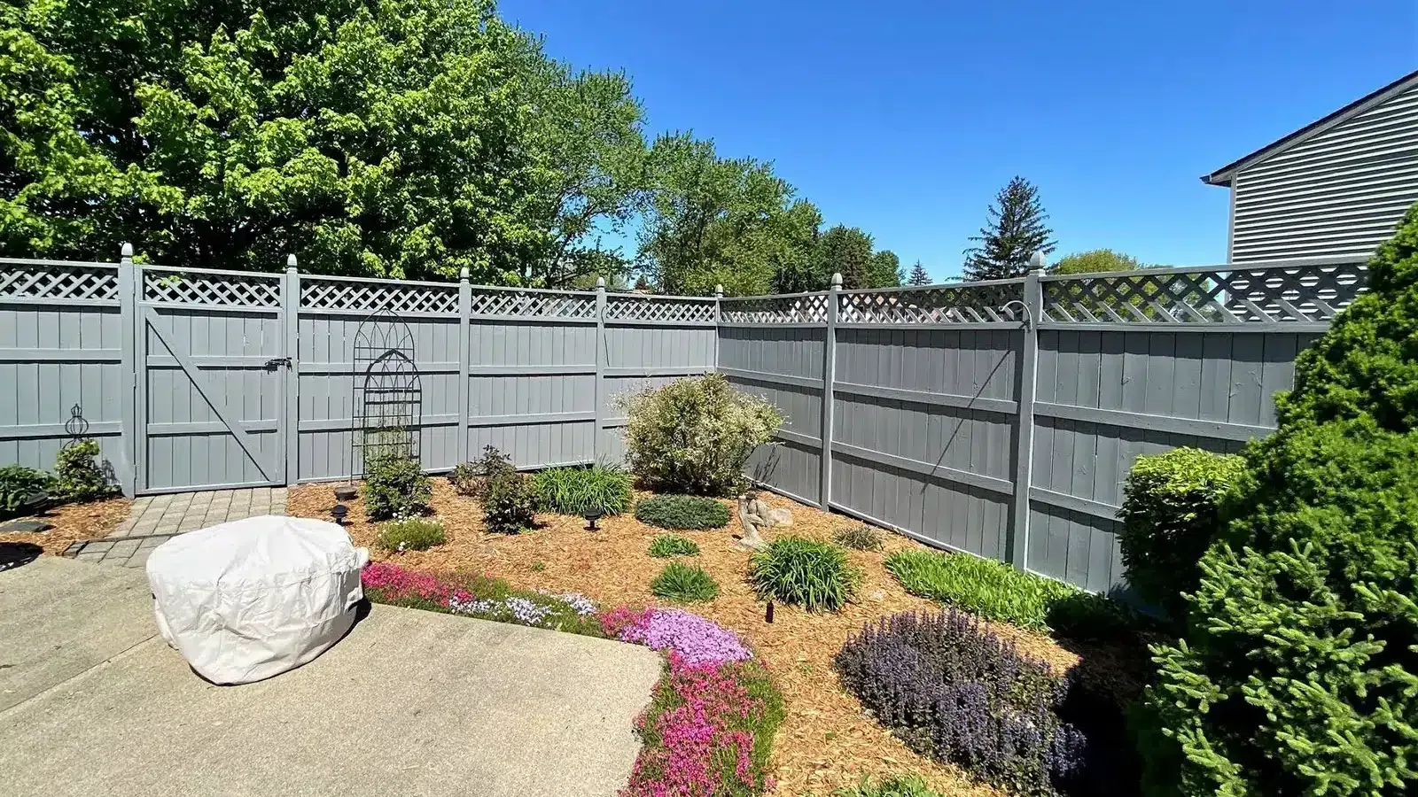 Gray wooden fence surrounds a small garden bed with colorful flowers and a covered white stone.