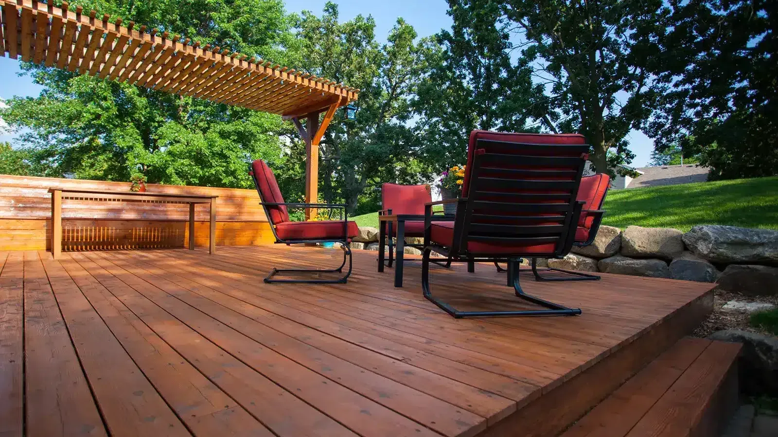 Wooden deck with red chairs, pergola, and green trees.