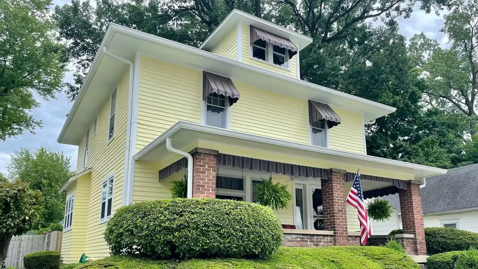 Yellow house with awnings, brick columns, porch, and American flag.