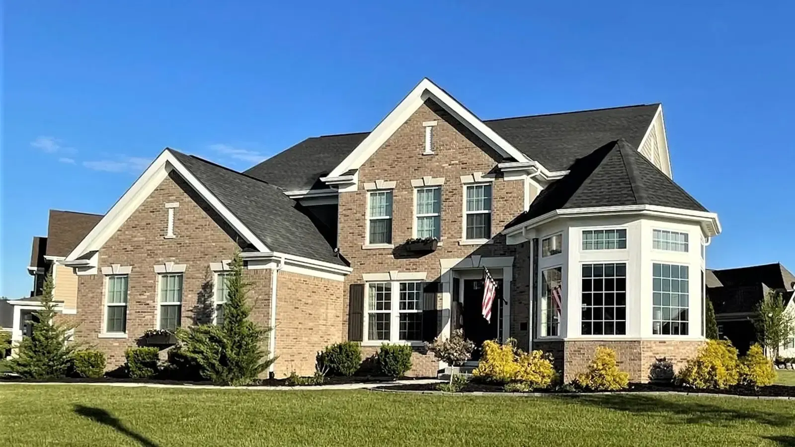 Two-story brick house with dark roof, bay window, and manicured lawn under a blue sky.