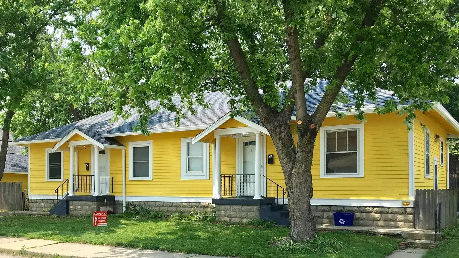 Yellow duplex with white trim, front porches, and a tree. Green grass and a blue recycling bin are visible.