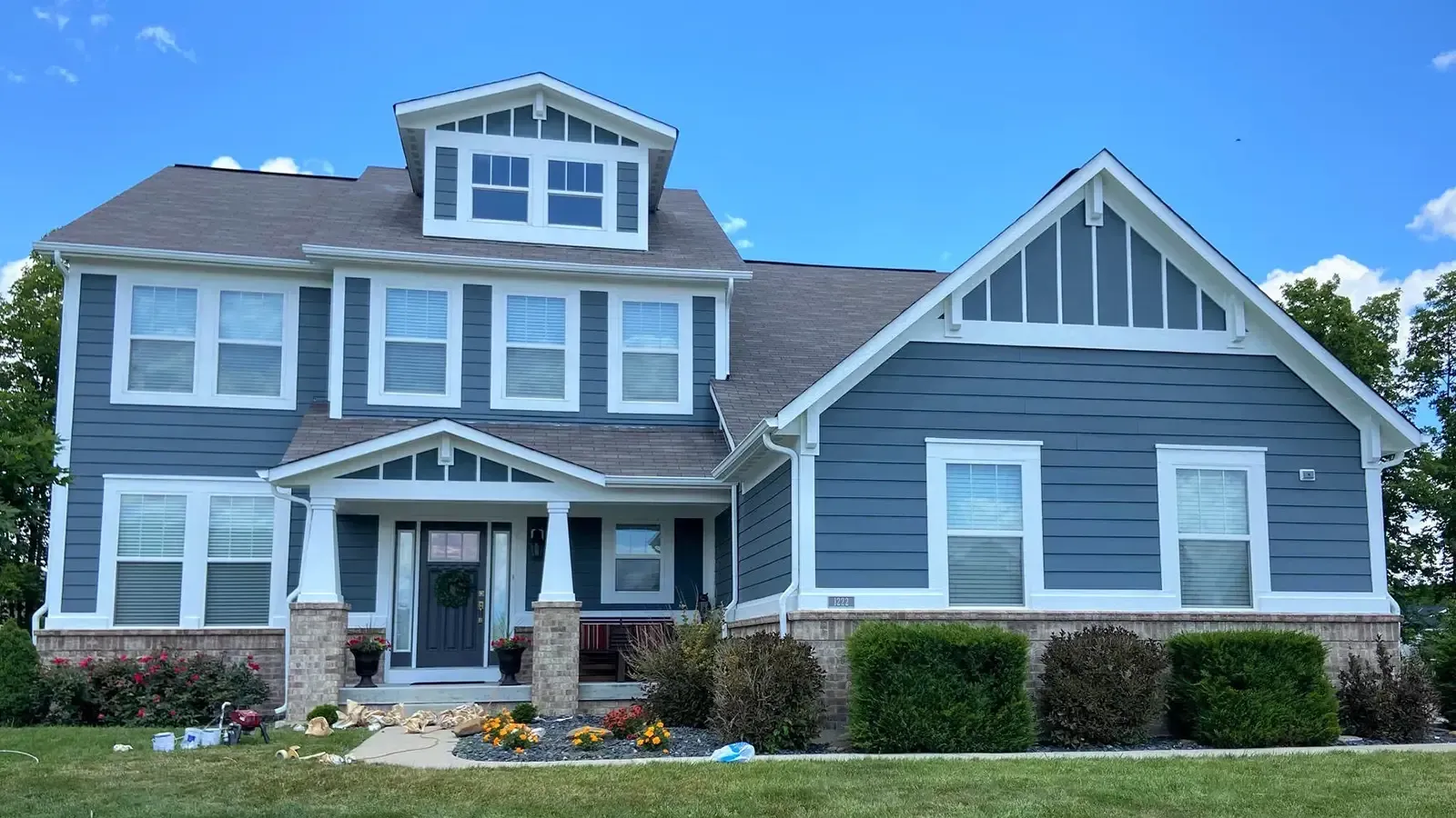 Two-story blue house with white trim and brick base, landscaped lawn under blue sky.