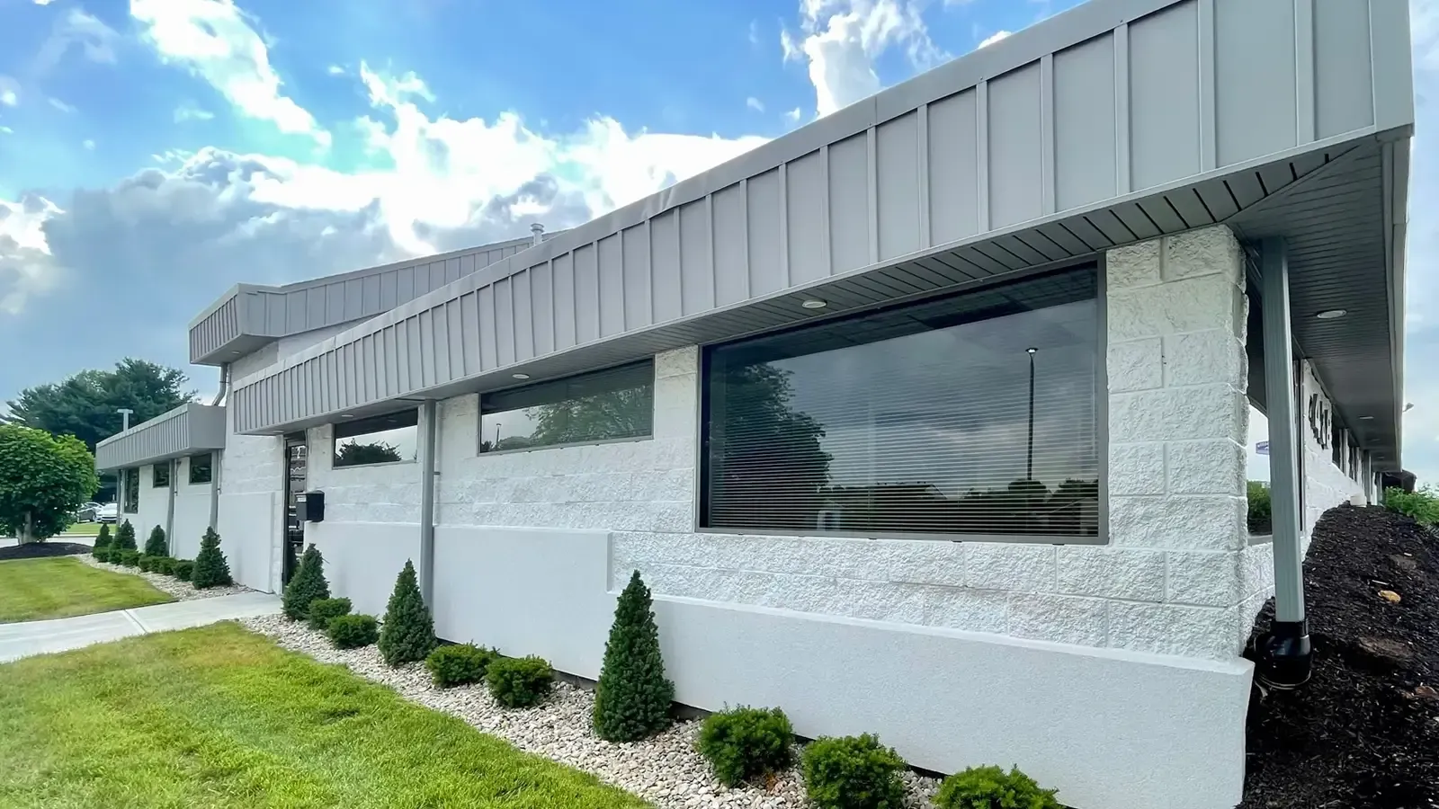 White building with gray roof and trim, large windows, green lawn, and shrubs.