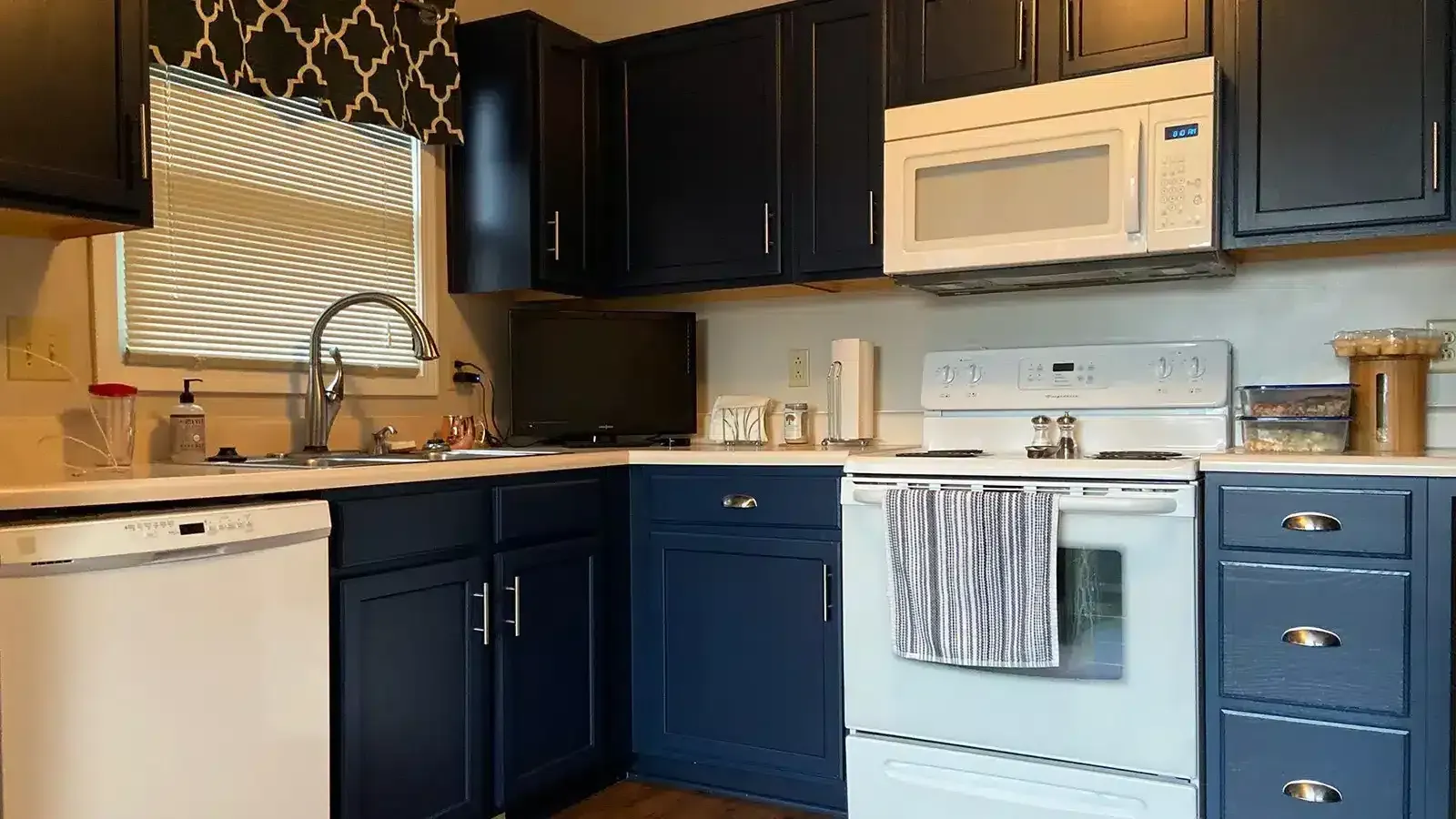 A kitchen with navy blue cabinets, white appliances, and a window with patterned shade.