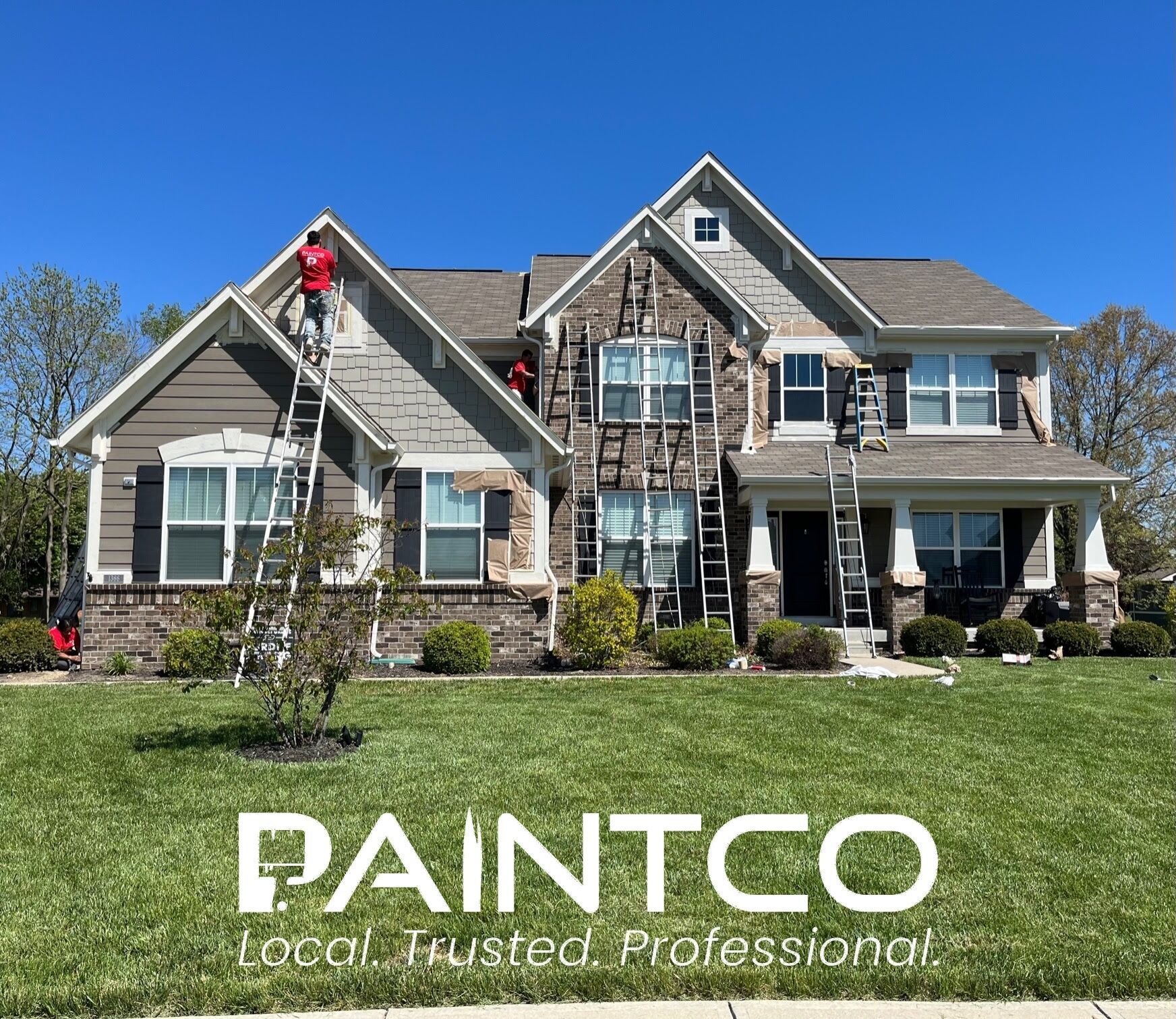 House being painted by PAINTCO. Workers on ladders. Brown siding, stone facade. Green grass, blue sky.