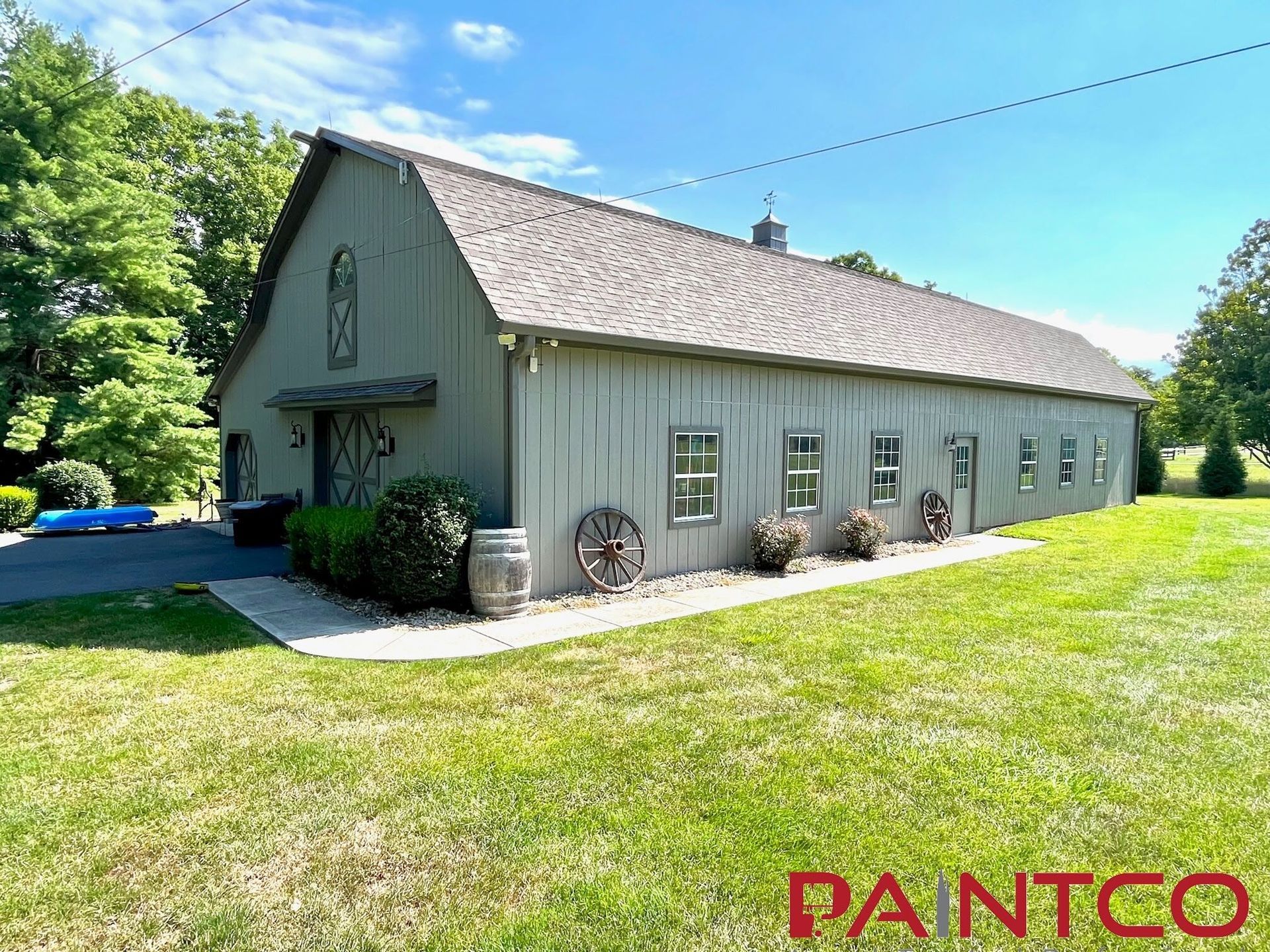Gray barn with a light-colored roof sits on a grassy lawn.  Small windows and antique wagon wheels decorate the side.