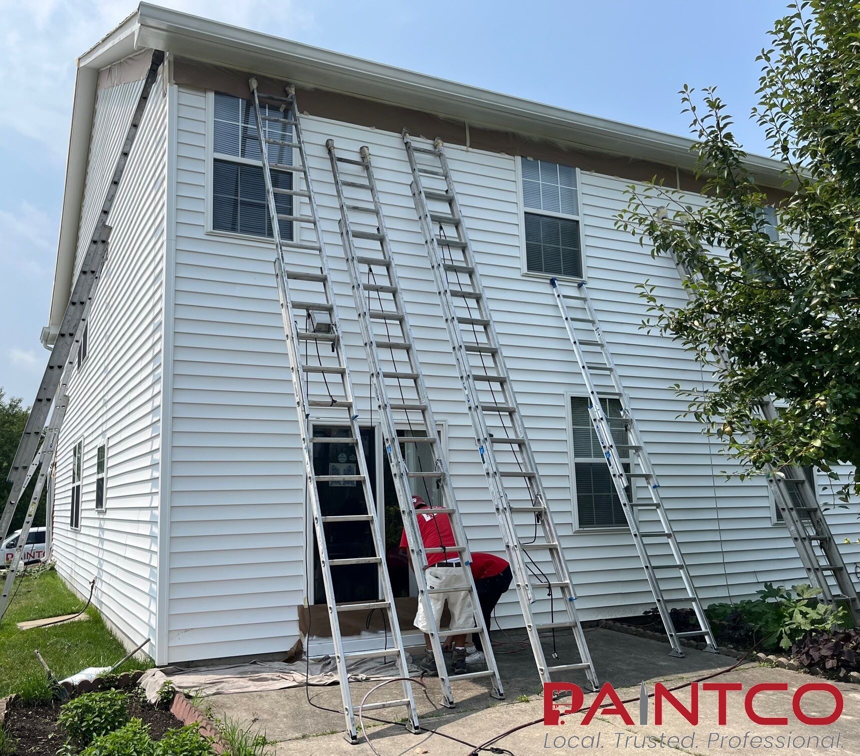 A two-story house being painted white by workers using multiple ladders on a sunny day.