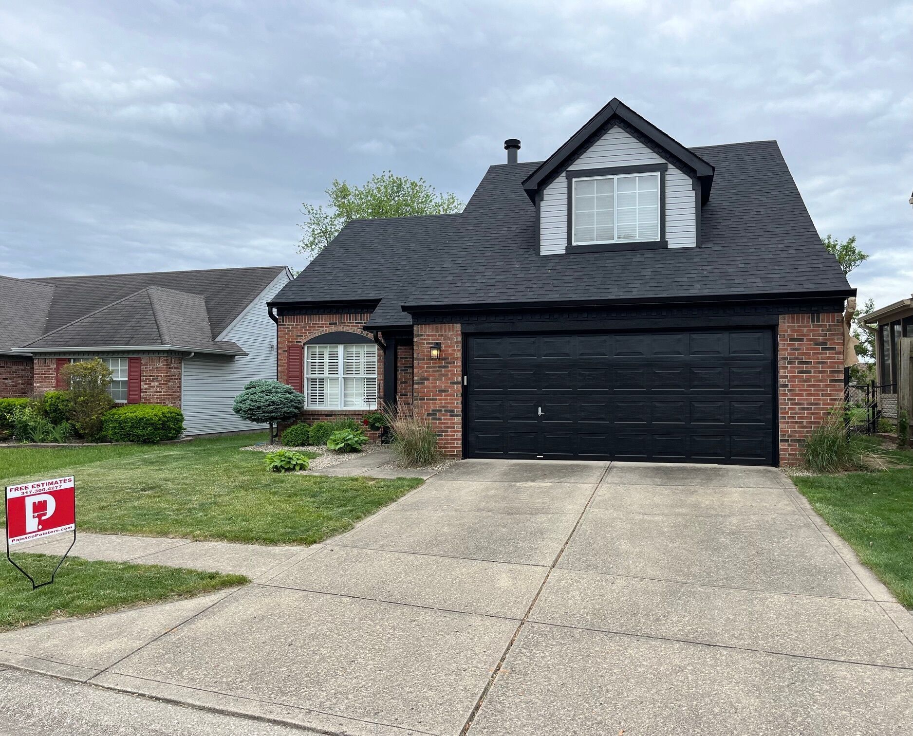 Brick house with black garage door and roof under a cloudy sky. A for sale sign is on the front lawn.