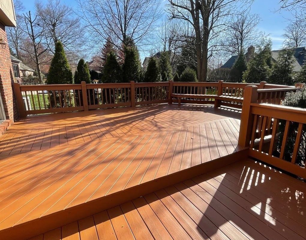Wooden deck with brown stain, railing, and surrounding trees under a sunny sky.