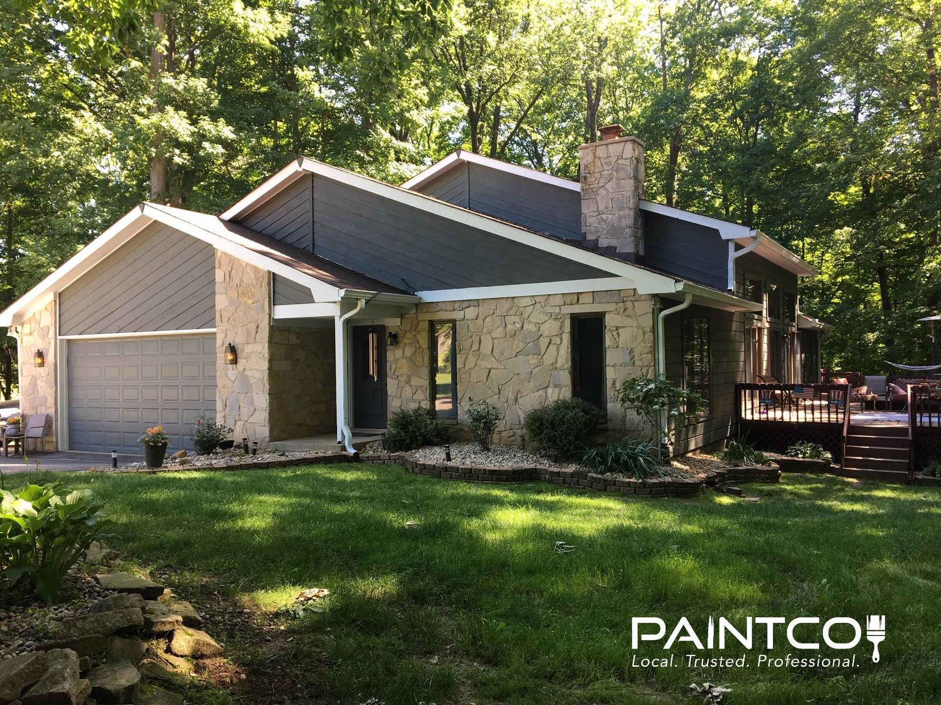 House with gray roof and garage, beige stone walls, brown chimney, and green yard.