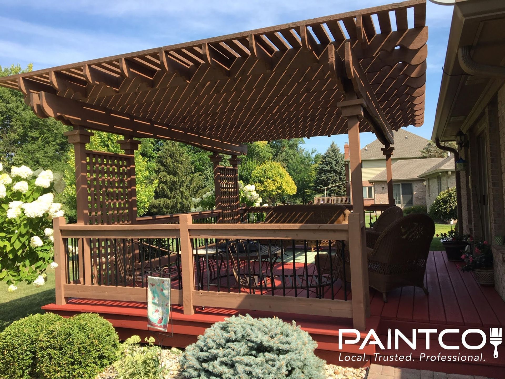 Brown-stained wooden pergola over a deck, with black railings, a brick house, and shrubbery.