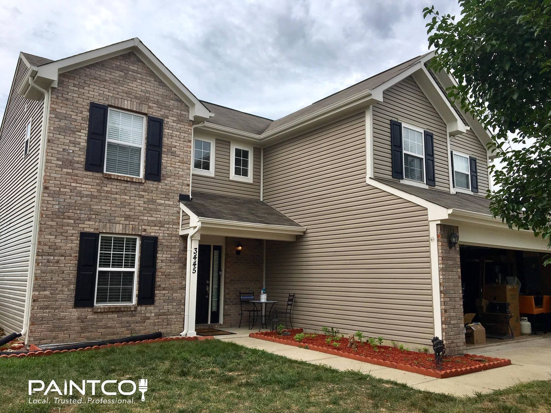 Two-story house with brick and tan siding, black shutters, brown roof, and red mulch landscaping.