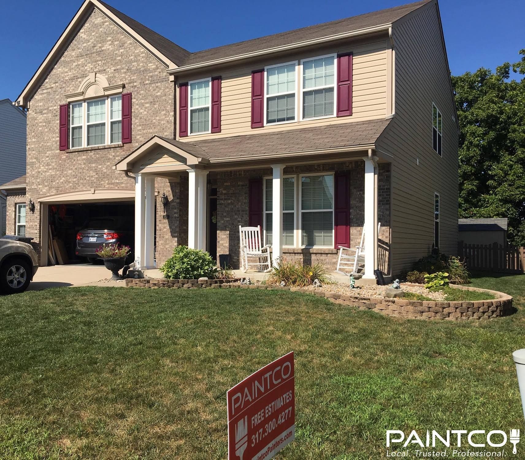 Two-story house with beige siding, brick, and burgundy shutters. A lawn and a PAINTCO sign are in the foreground.