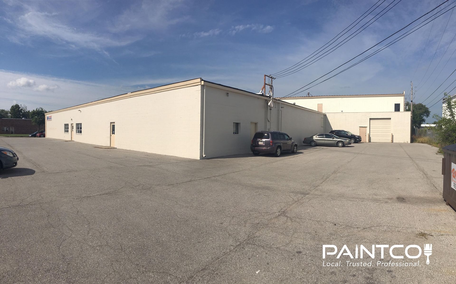 Exterior view of a light-colored commercial building with parked cars on a paved lot under a blue sky.